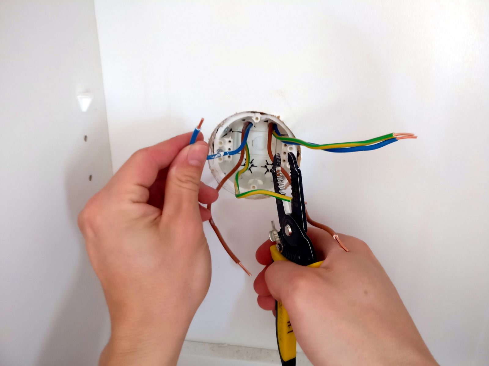 Hands stripping a wire with pliers at an open junction box on a white wall