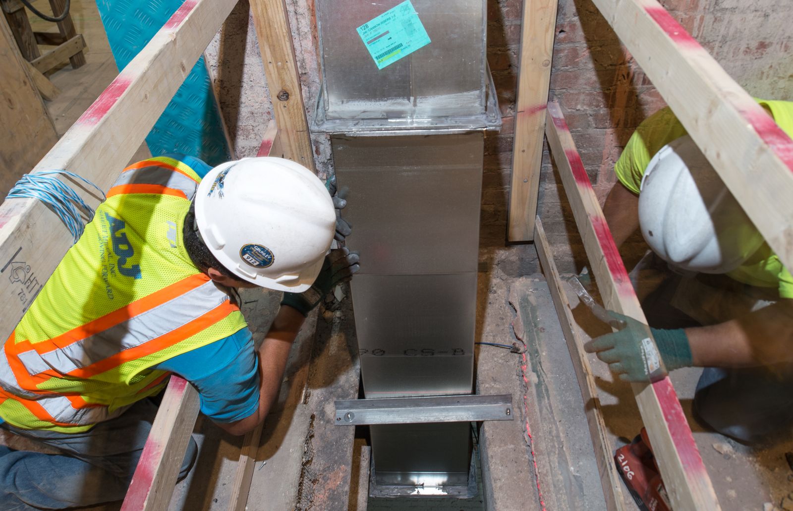 Two workers in hard hats fitting a rectangular duct between floor framing, seen from above